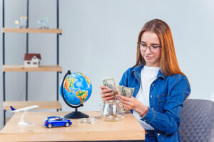 Young girl counts money from a piggy bank while sitting at a table. Concept of financial literacy and savings. Transparent piggy bank with banknotes.